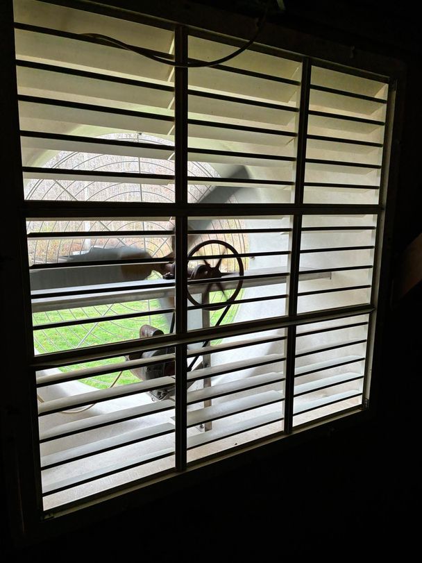 Cleaned white barn fan shutter back in place, backlit by daylight with the belt-driven exhaust fan running behind