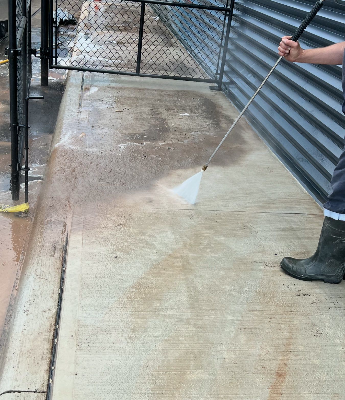 A new dealership walkway mid-wash: construction mud still on the far half, the near half washed back to clean concrete, a wand spraying at the wash line beside metal cladding