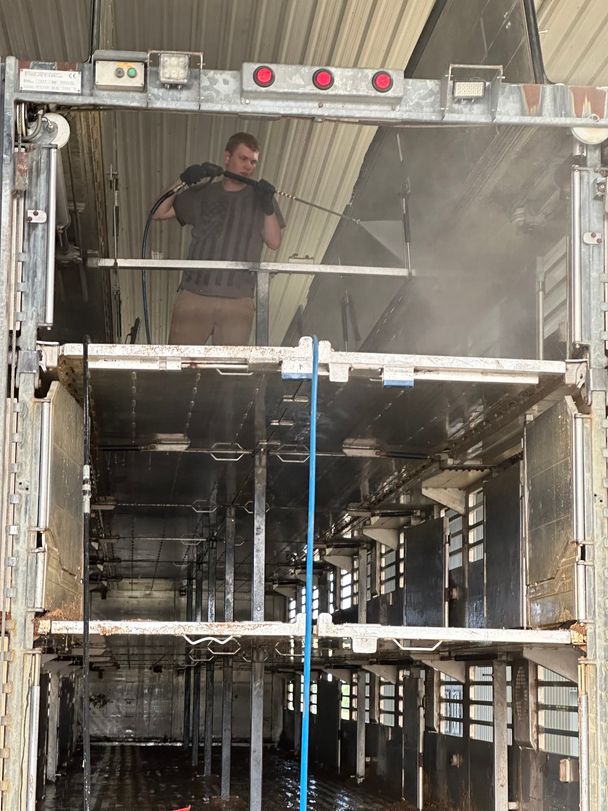 Crew member pressure washing the top deck of a triple-deck hog trailer inside a truck wash bay