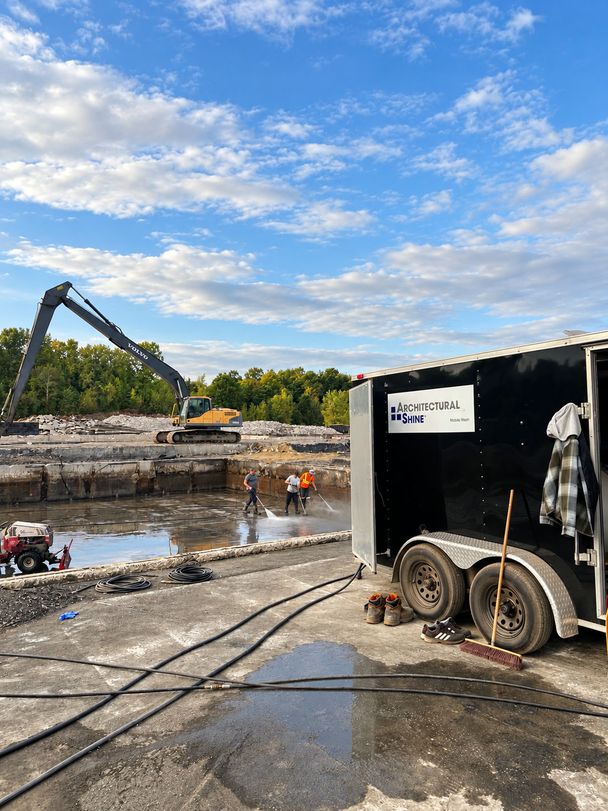 Three crew members pressure-washing the floor of a large emptied concrete manure pit under a blue sky, a long-reach excavator beyond and the Architectural Shine mobile wash trailer with hoses in the foreground