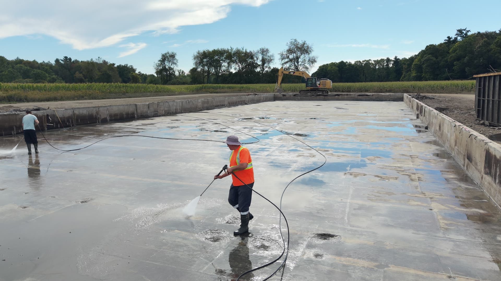 Crew pressure washing the wet floor of an emptied concrete pit, an excavator and a cornfield beyond