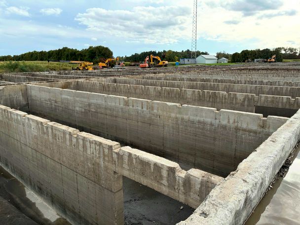 Rows of washed grey concrete manure storage cells under a cloudy sky, excavators parked beyond