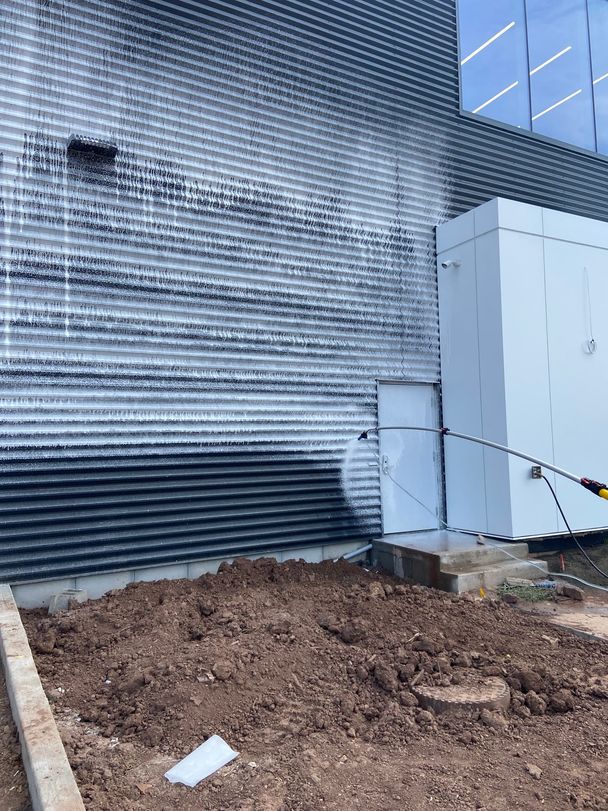 White cleaning foam running down dark ribbed metal cladding as a wand rinses a new commercial building wall