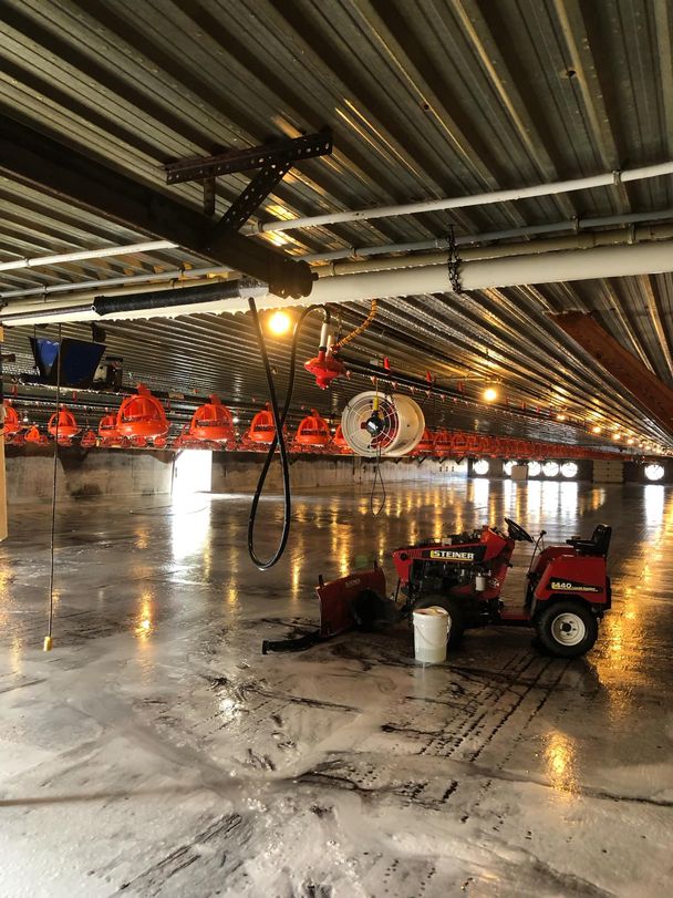 Compact barn tractor with scraper blade on a foam-streaked poultry barn floor during wash, pan feeders raised