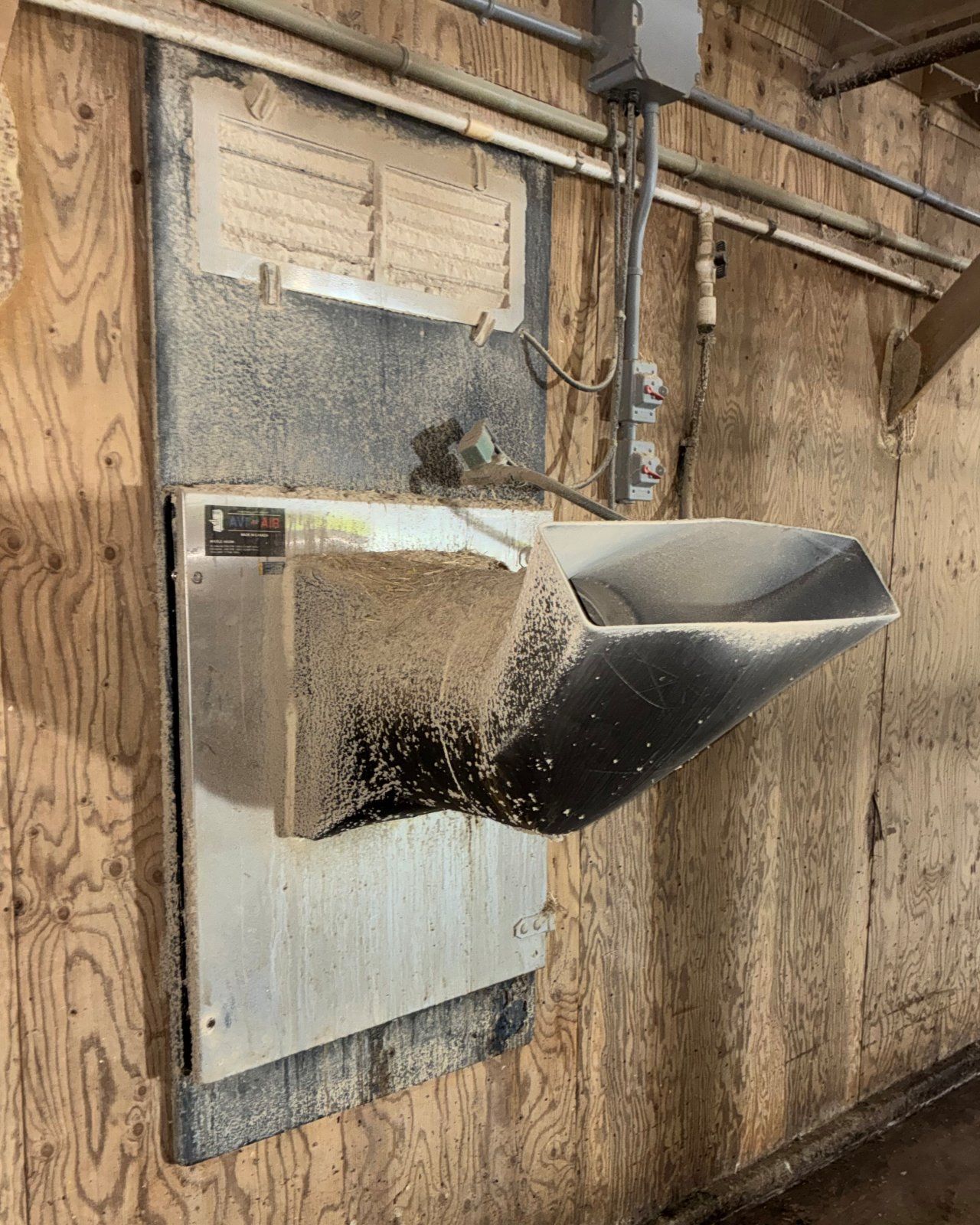 Barn heater on a plywood wall in a poultry barn before cleaning: louvres packed with dust and a dust-coated housing and hood.