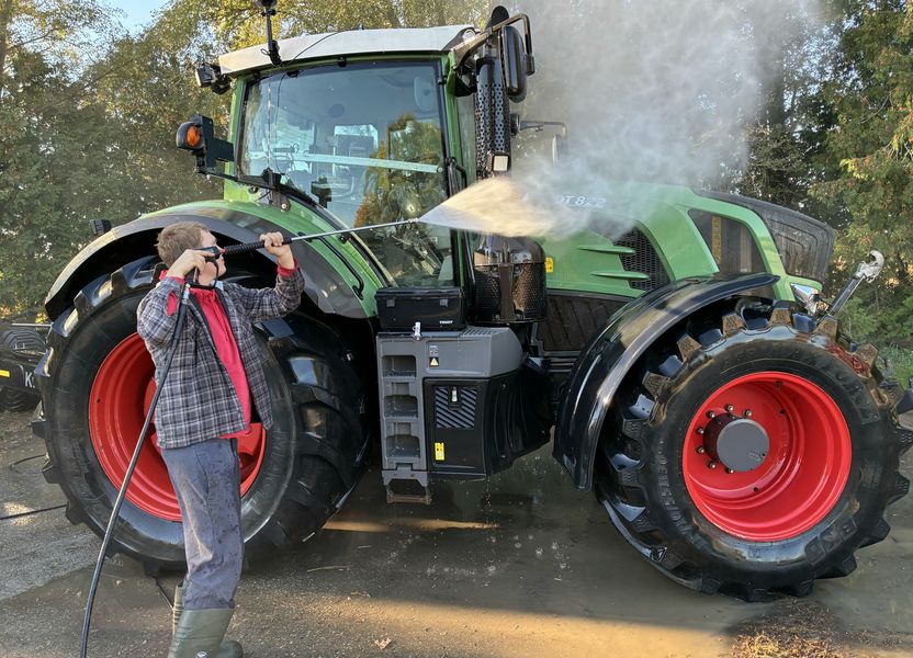 A crew member pressure-washing the side of a green tractor with red wheels, spray catching the morning sun
